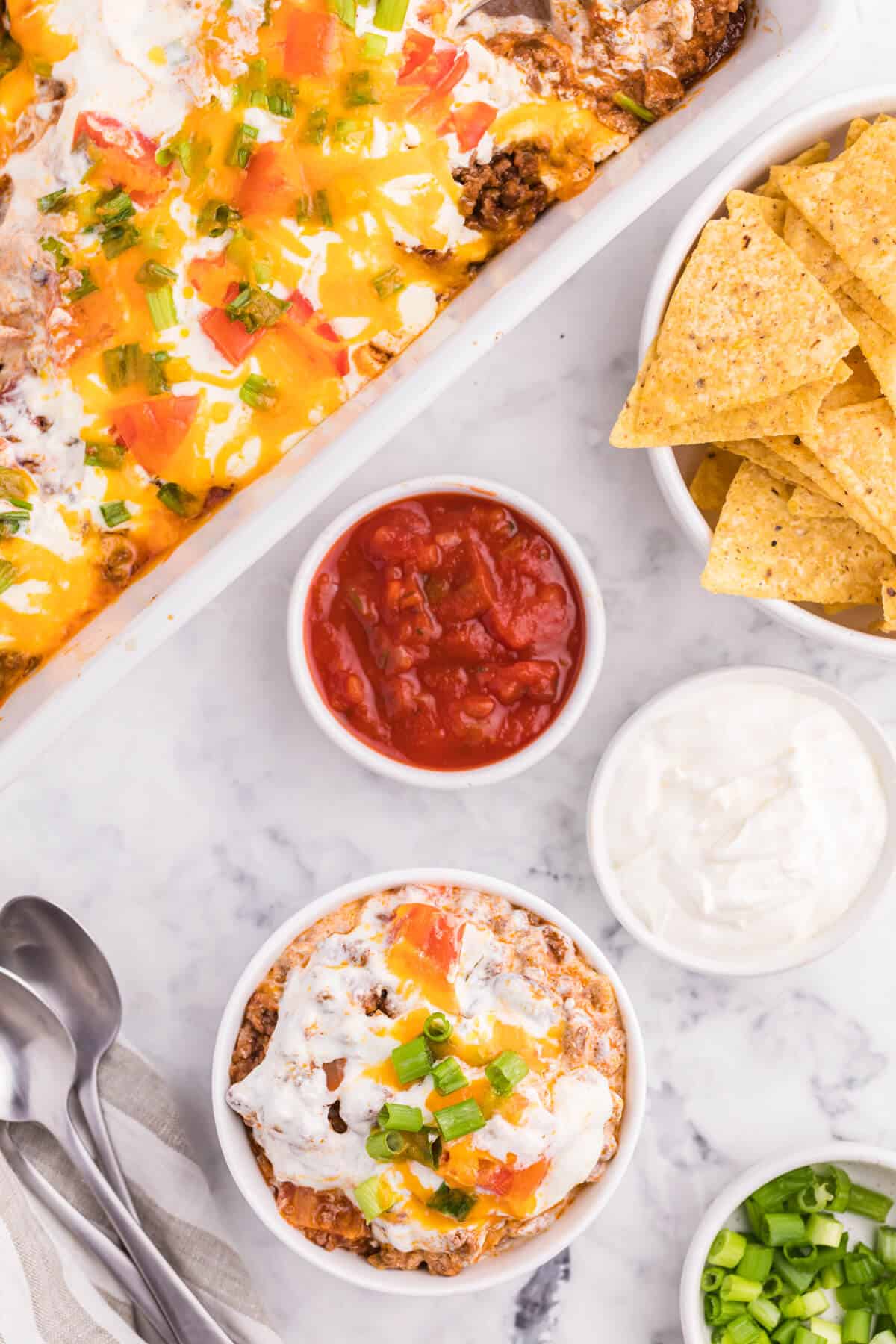 A bowl of taco casserole with toppings around it.