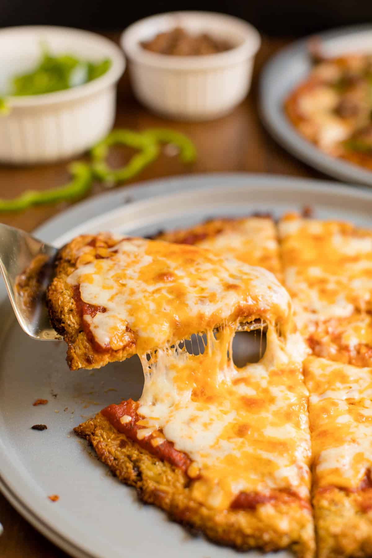 A spatula lifting a slice of cheese pizza made with a cauliflower crust off a pizza pan.