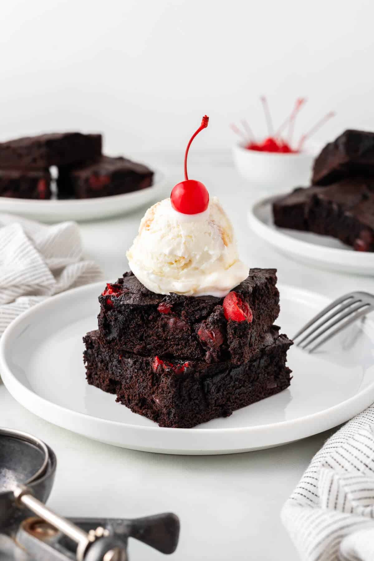 Two cherry chocolate brownies stacked on a plate topped with ice cream and a cherry.