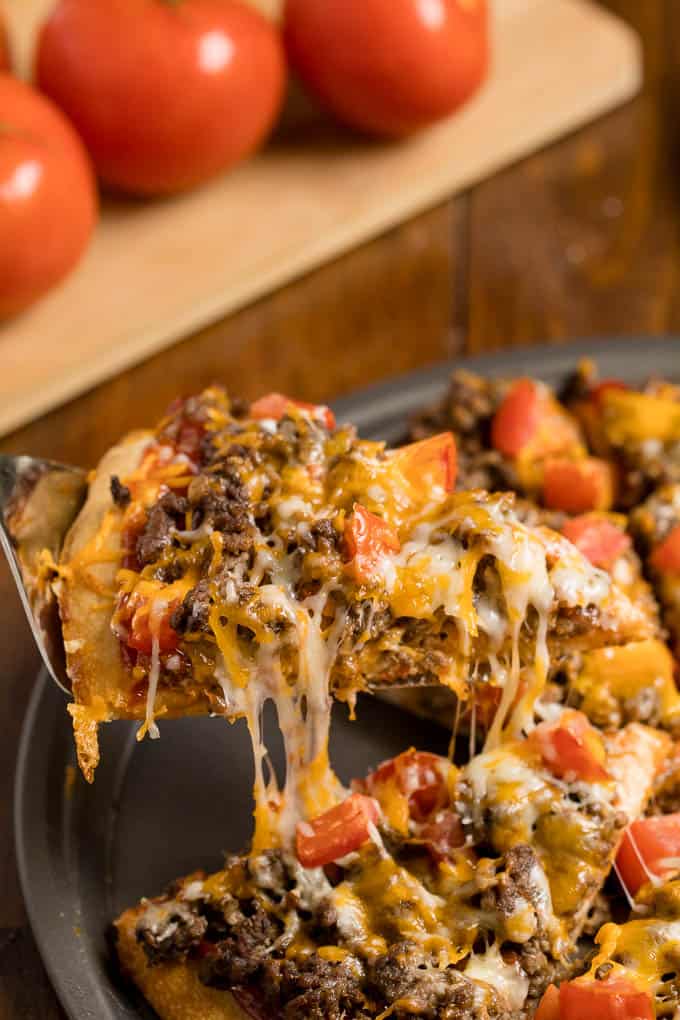 A serving spatula lifting a slice of cheeseburger pizza off a pizza pan. 