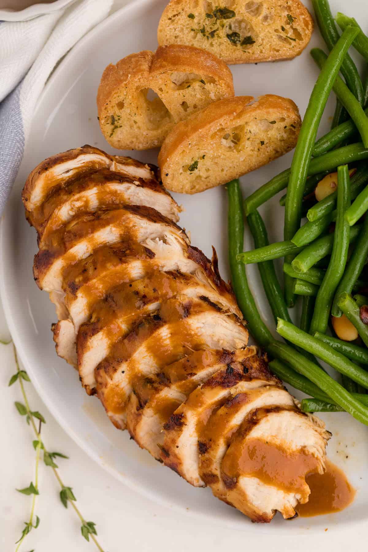 A sliced grilled herb chicken breast on a plate with garlic bread and green beans.