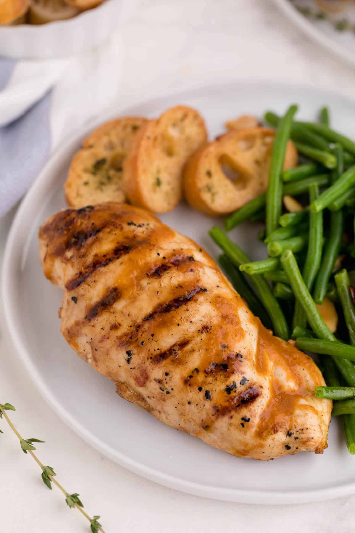A grilled herb chicken breast on a dinner plate with slices of garlic bread and green beans.