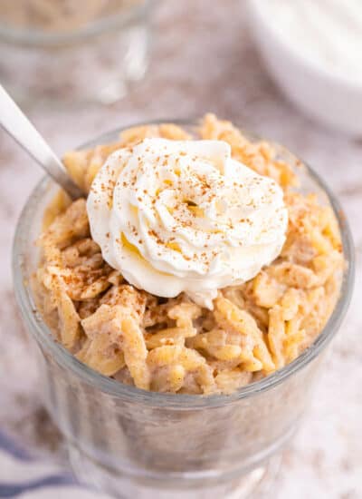 Orzo pudding topped with whipped cream in a parfait dish with a spoon.