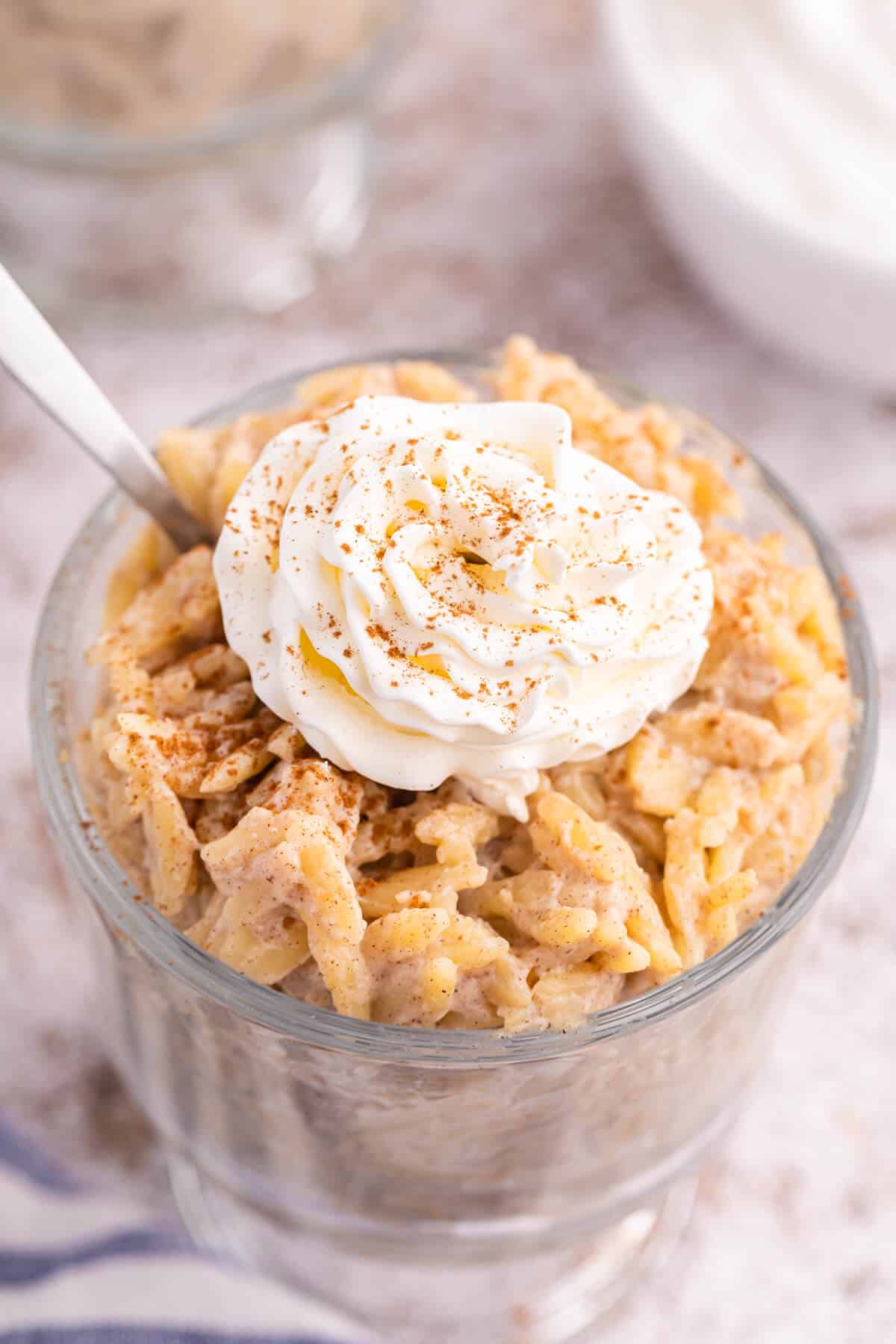 Orzo pudding topped with whipped cream in a parfait dish with a spoon.