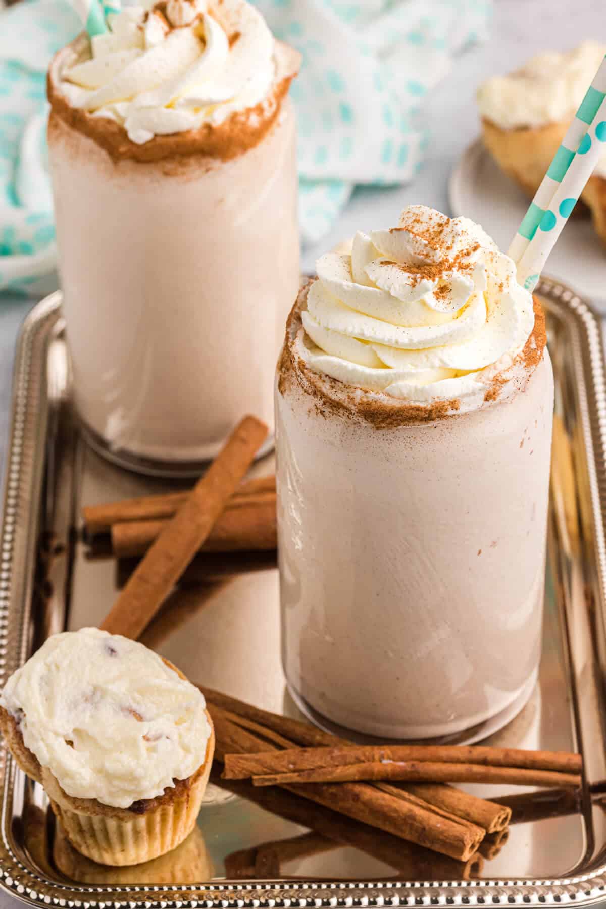 Two cinnamon roll coffee milkshakes on a tray with a cinnamon roll and cinnamon sticks.