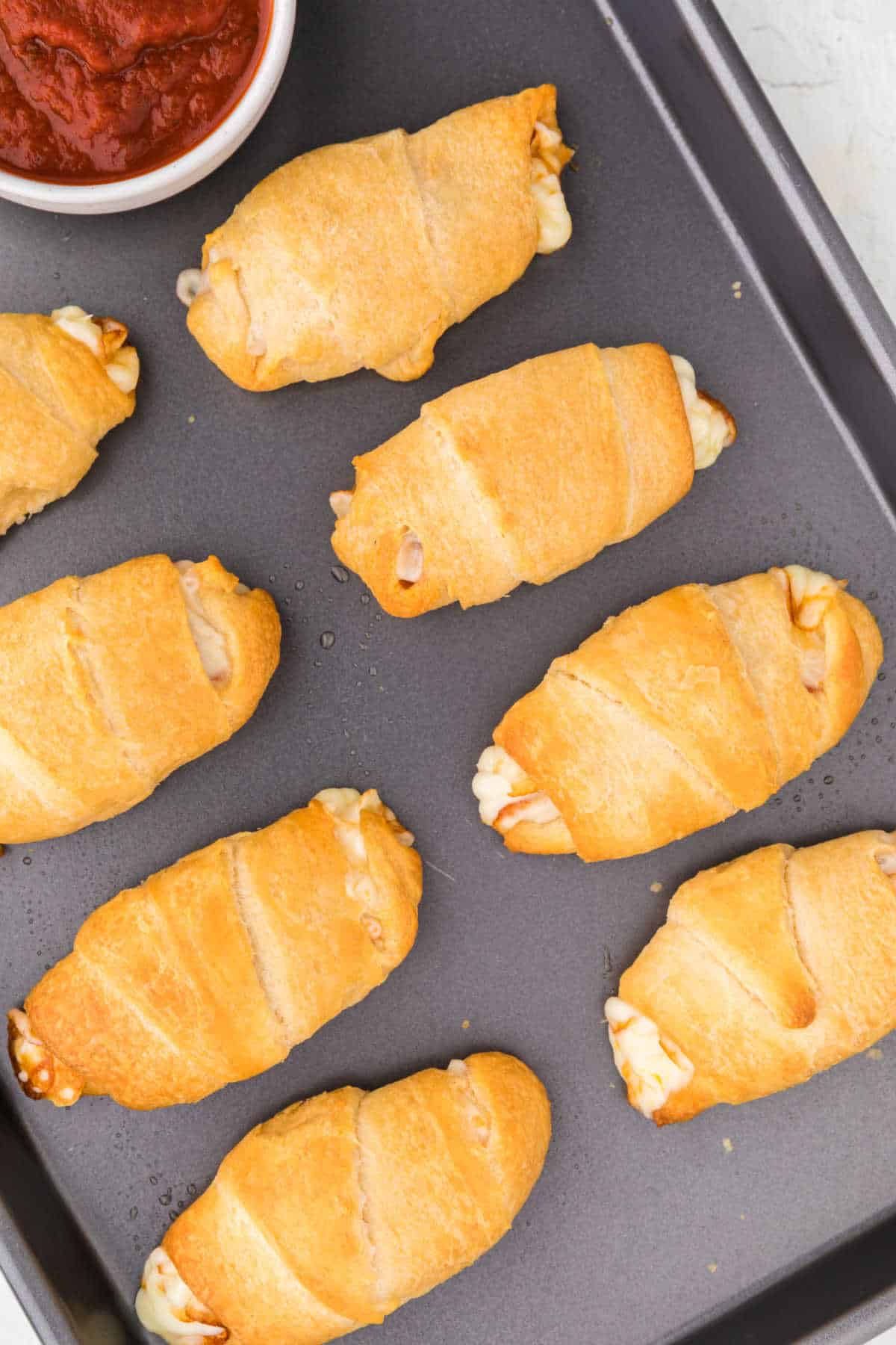 Pizza rolls on a sheet pan with a bowl of pizza sauce.