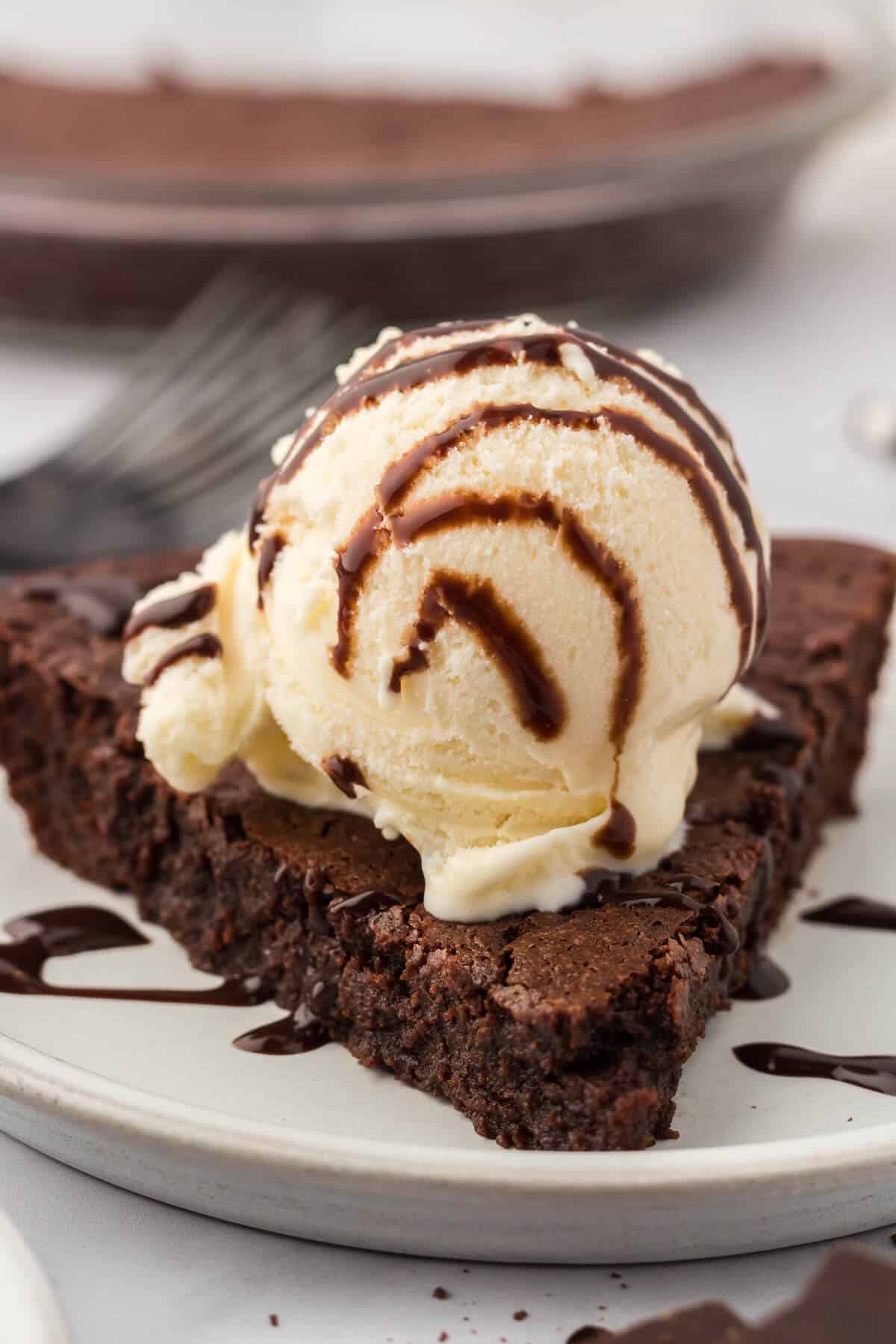 A slice of fudge pie on a plate topped with vanilla ice cream and chocolate sauce.