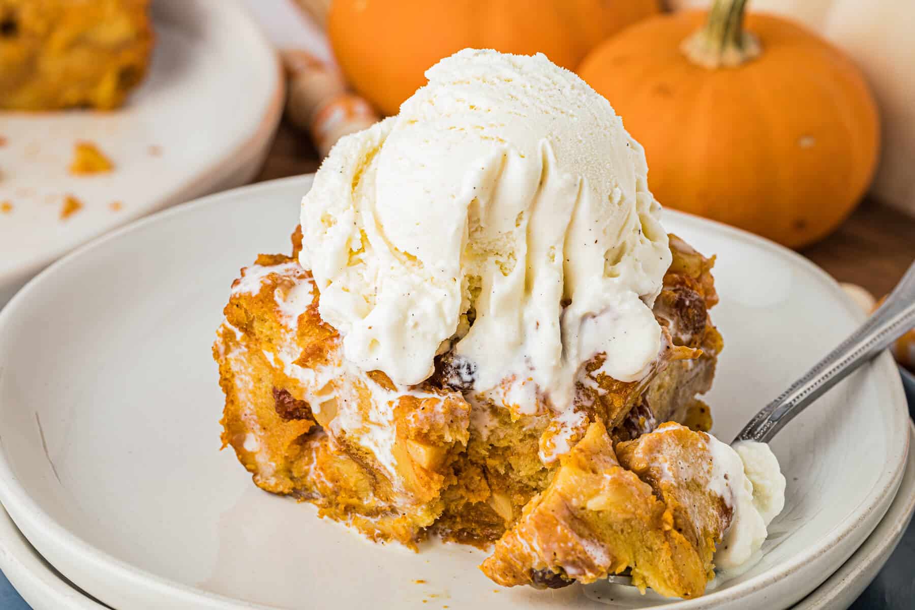 A piece of pumpkin bread pudding topped with vanilla ice cream on a plate with a fork.