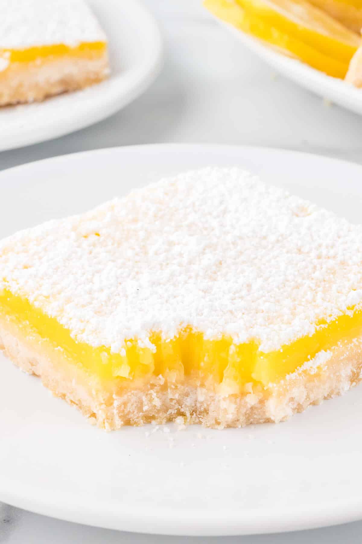 A lemon bar on a plate with a bite out of it.