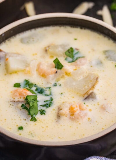 Bowl of creamy shrimp chowder with potatoes, shrimp, and chopped parsley, with fresh parsley and shredded cheese nearby.