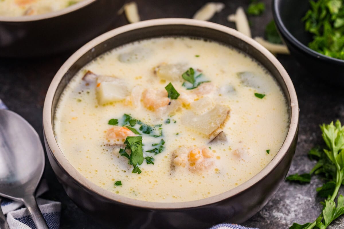Bowl of creamy shrimp chowder with potatoes, shrimp, and chopped parsley, with fresh parsley and shredded cheese nearby.