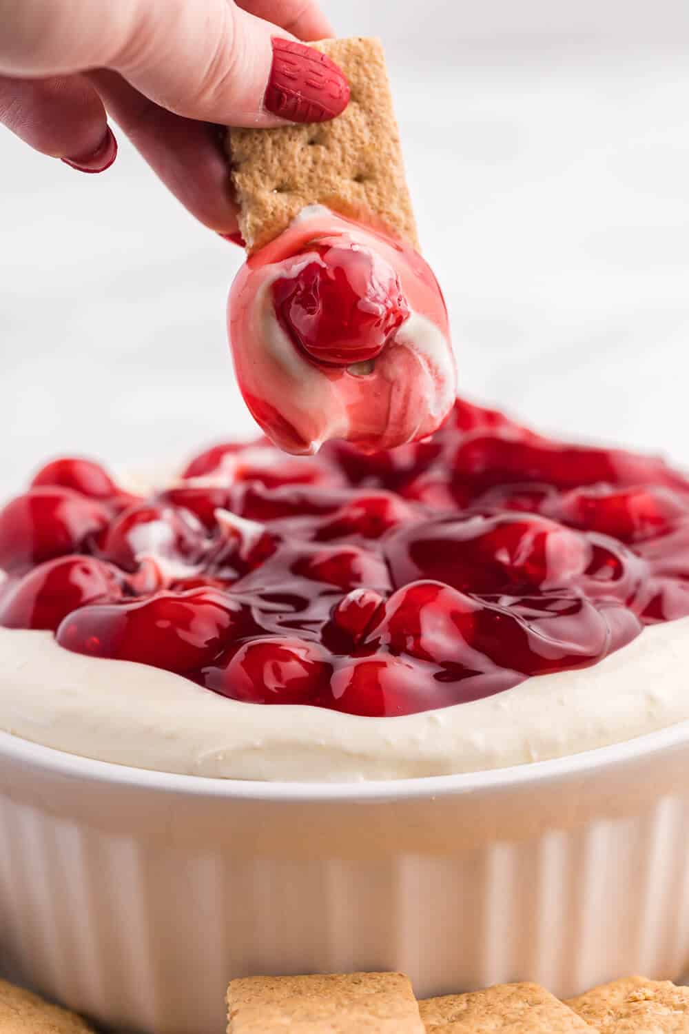 a hand dipping a graham cracker in cherry cheesecake dip