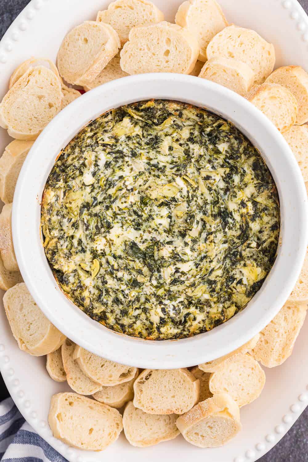 A bowl of spinach artichoke dip with slices of baguette.