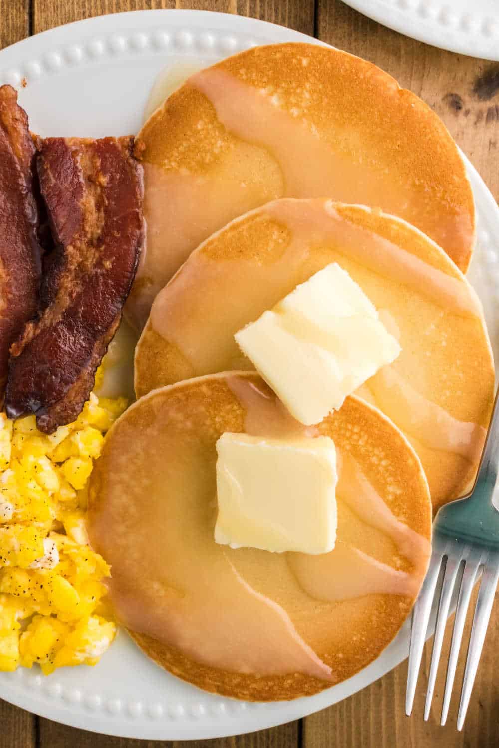 A plate of pancakes with pancake syrup, bacon and scrambled eggs.