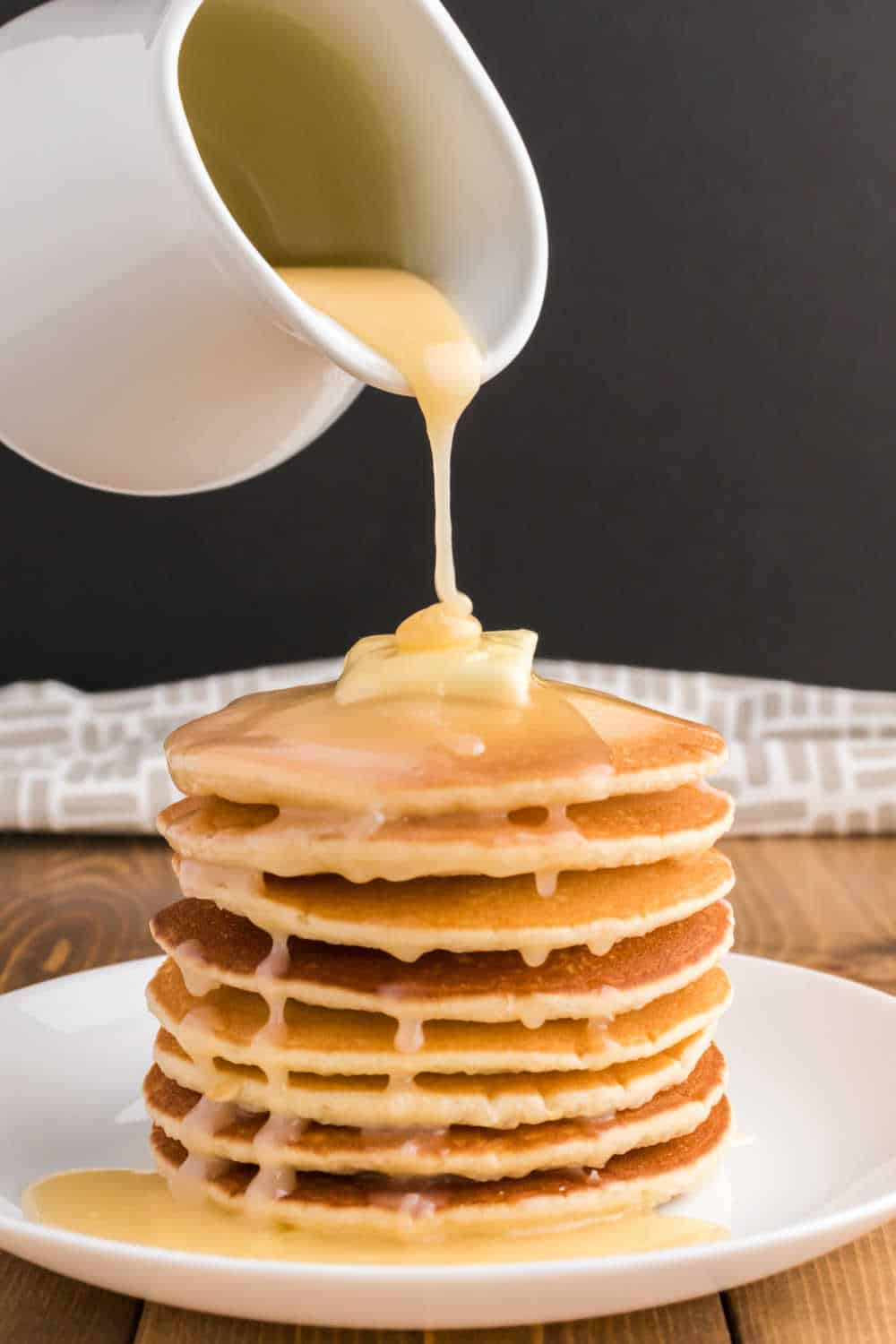 A pitcher drizzling pancake syrup over a stack of pancakes.