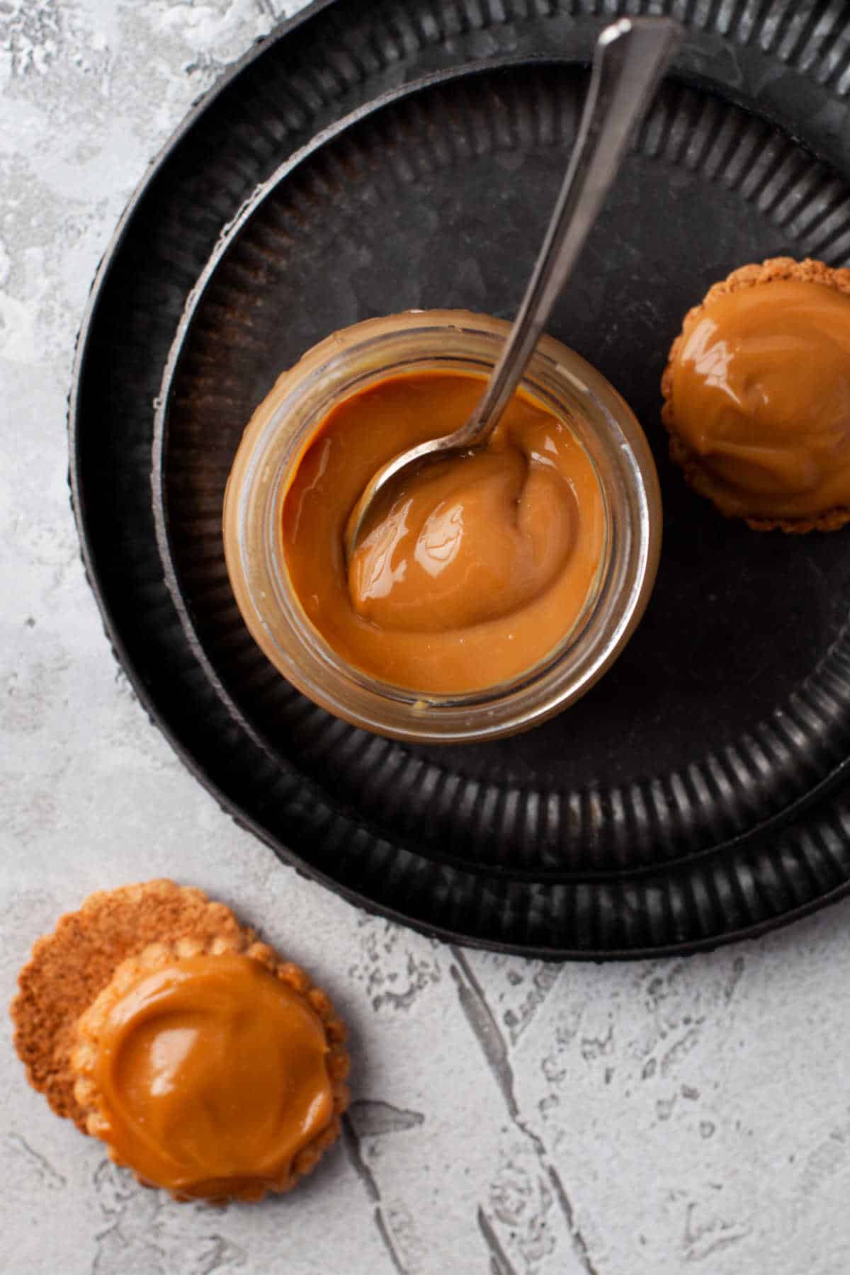 A jar of dulce de leche with a spoon on a platter with some cookies.