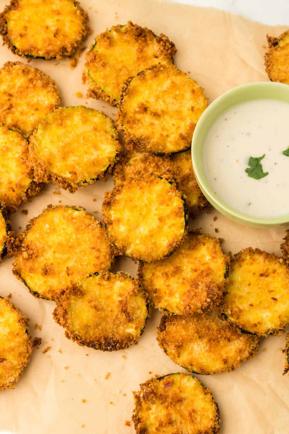 Air fryer zucchini chips on parchment paper with dipping sauce.