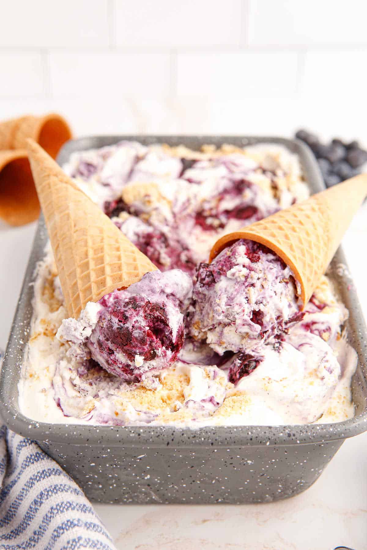 Two ice cream cones in a pan of blueberry pie ice cream.