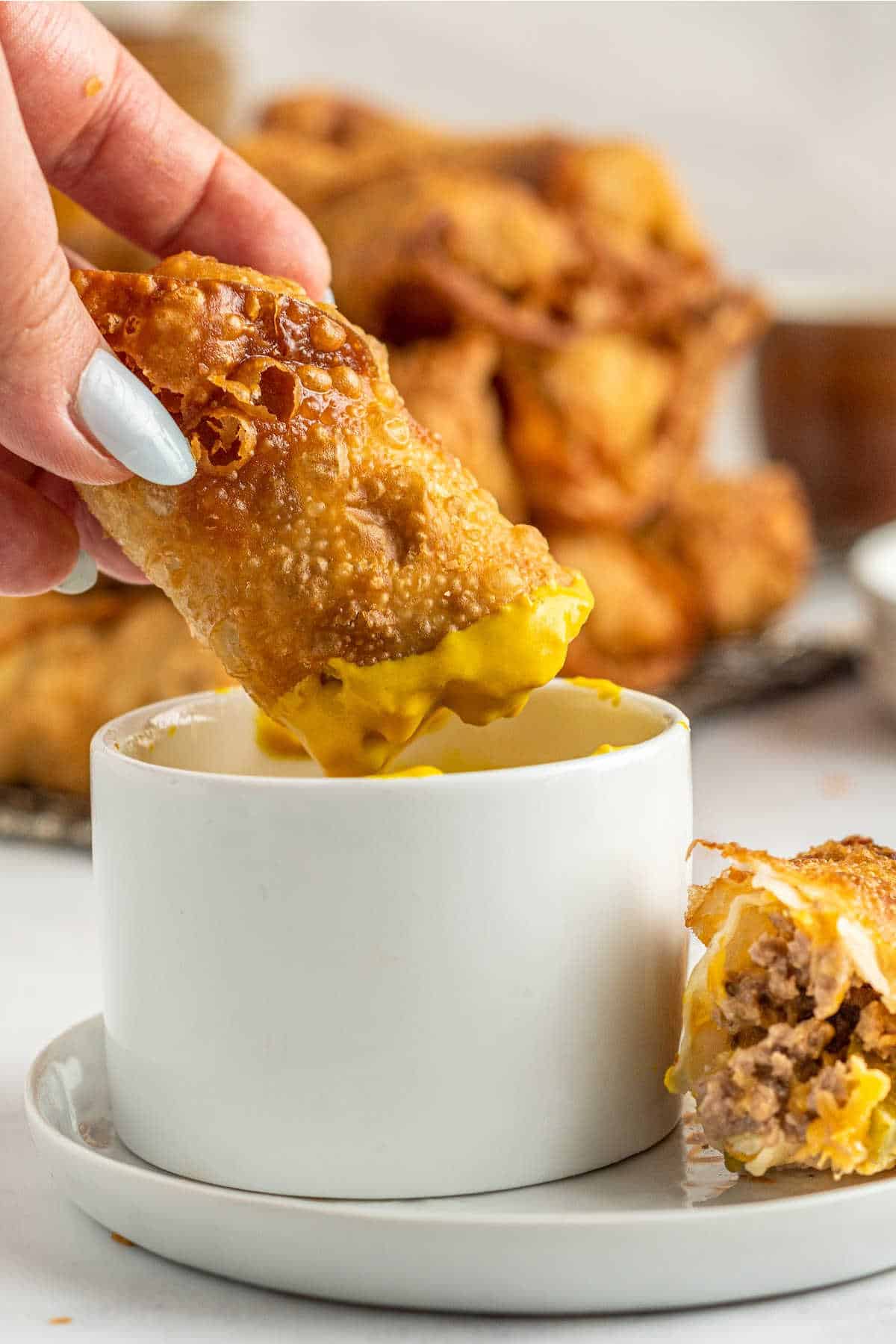 A hand dipping a cheeseburger egg roll in a dish of mustard.