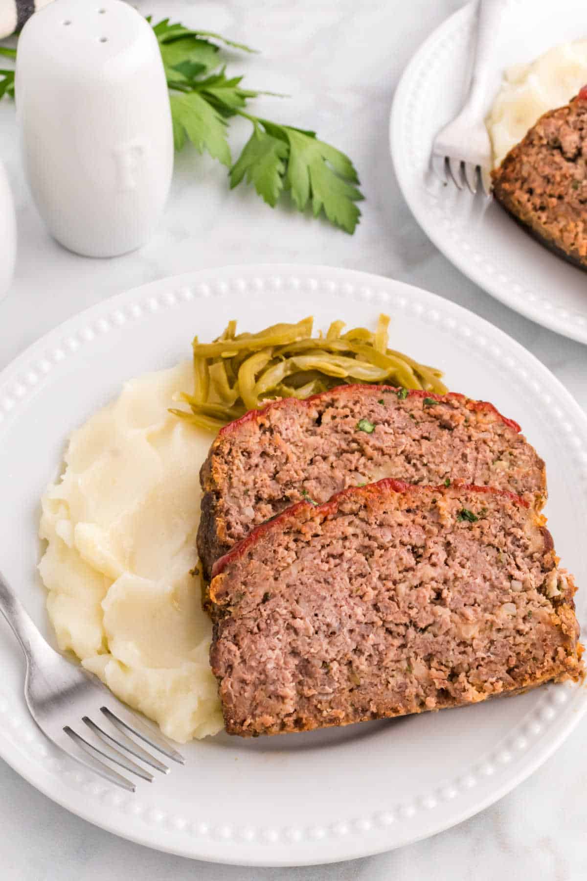 Slices of meatloaf with mashed potatoes and green beans on a plate with a fork.