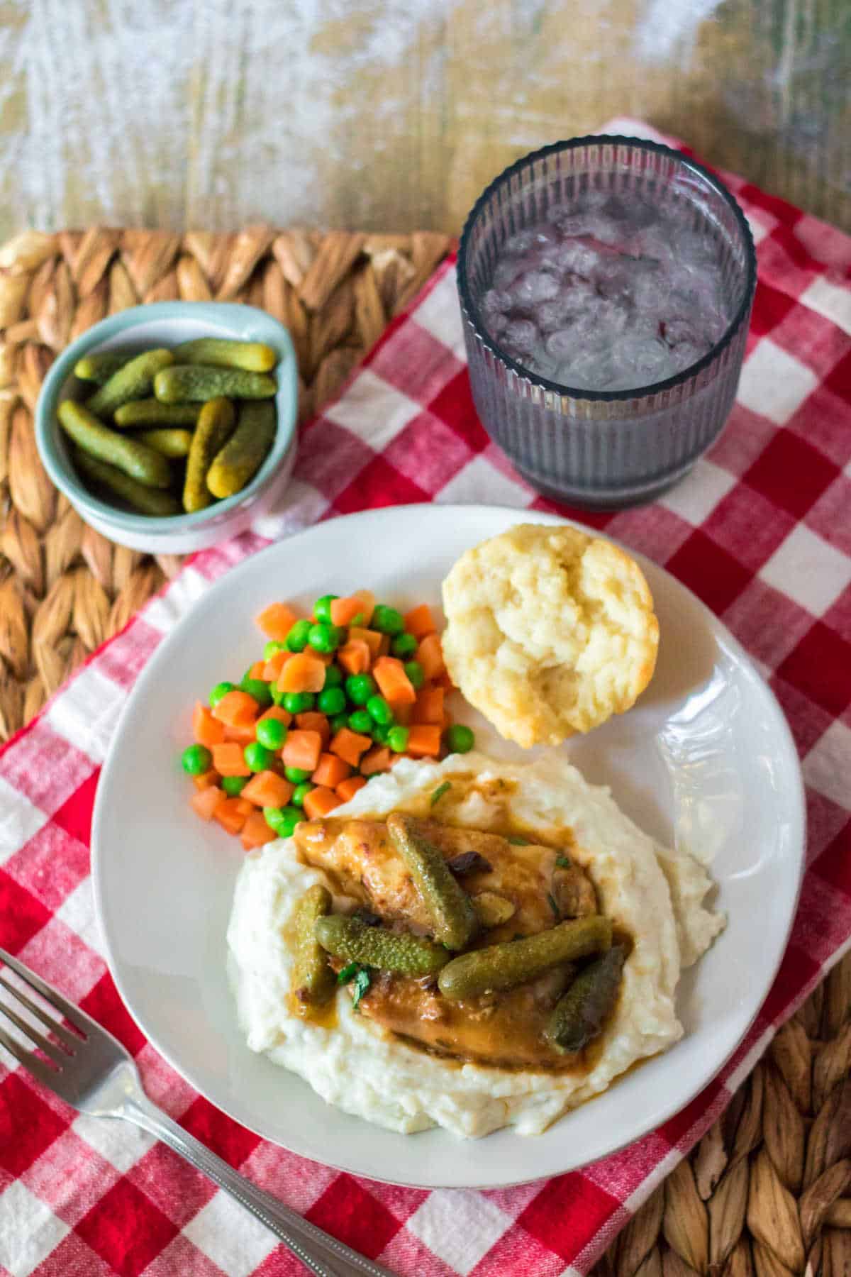 A plate with pickle chicken, mashed potatoes, peas, carrots and a bun.