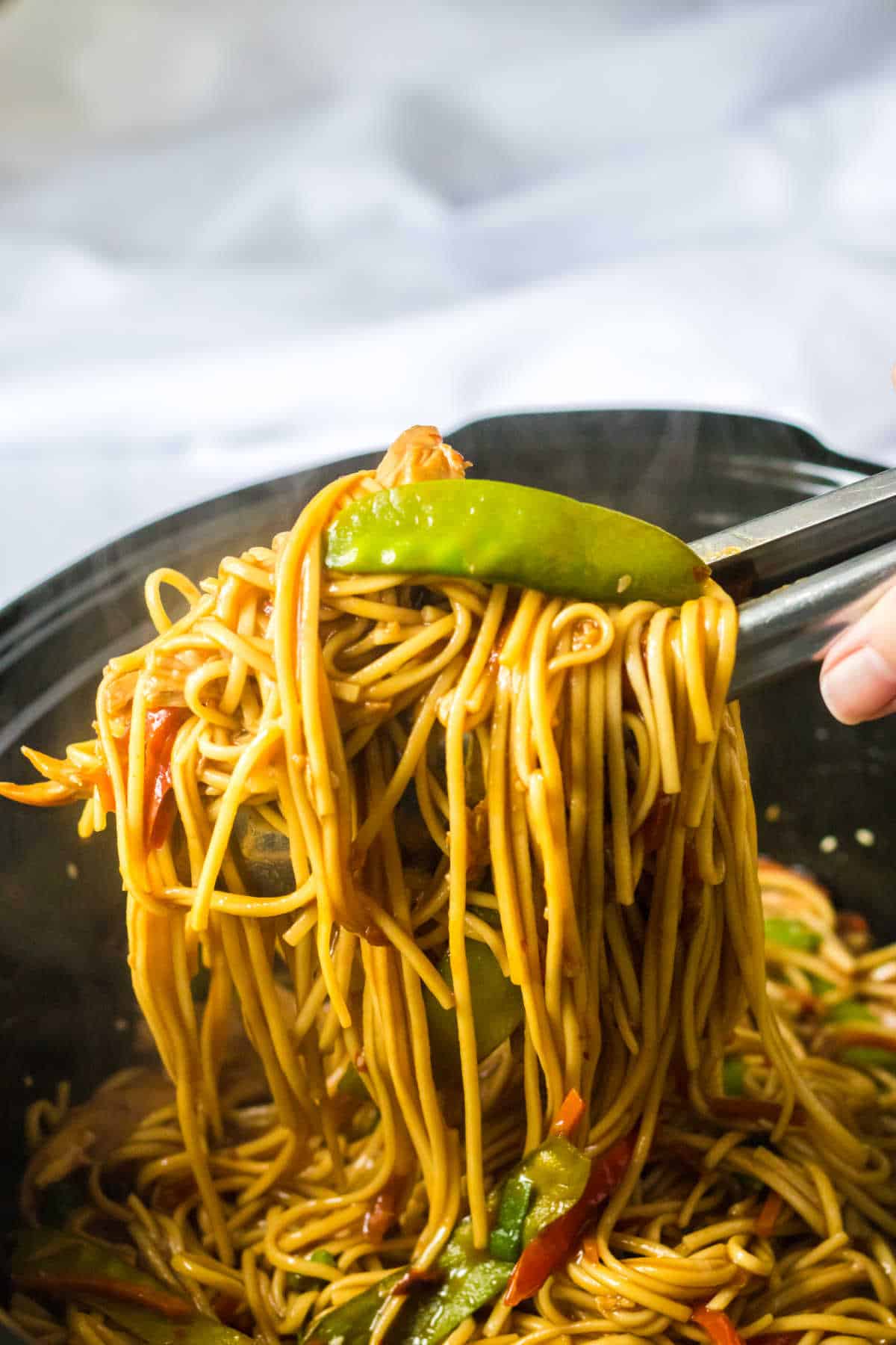 Tongs lifting chicken lo mein from a slow cooker.