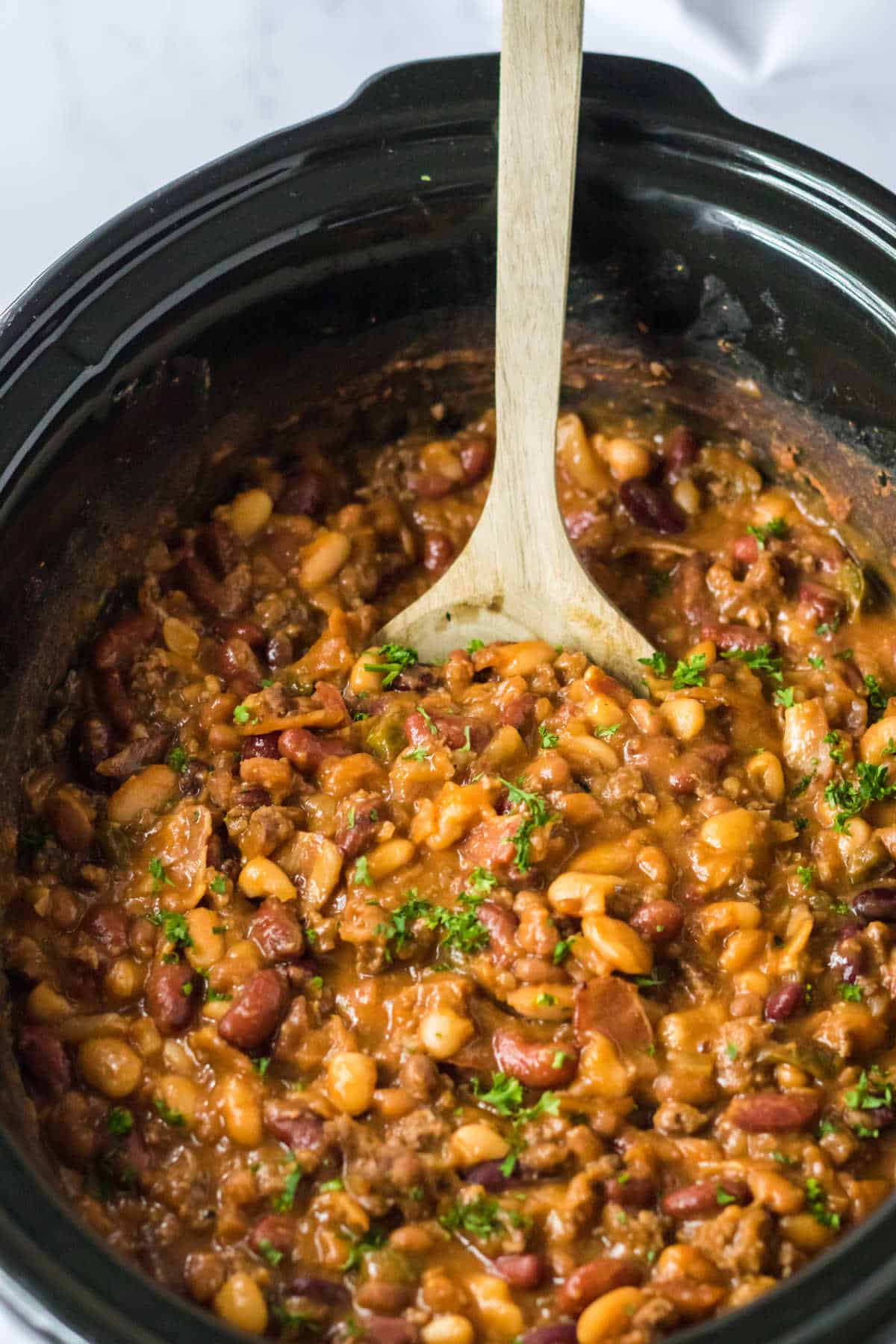 Cowboy beans in a slow cooker with a  wooden spoon.