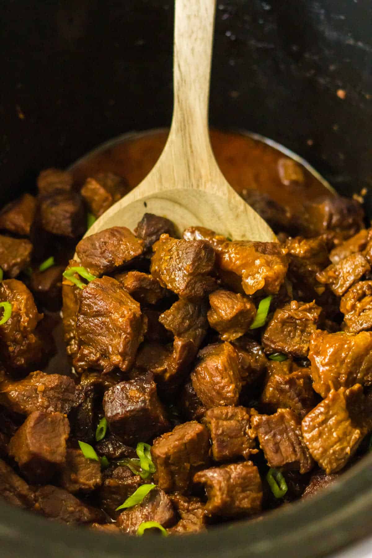 Honey Bourbon Steak Bites in a slow cooker with a wooden spoon.