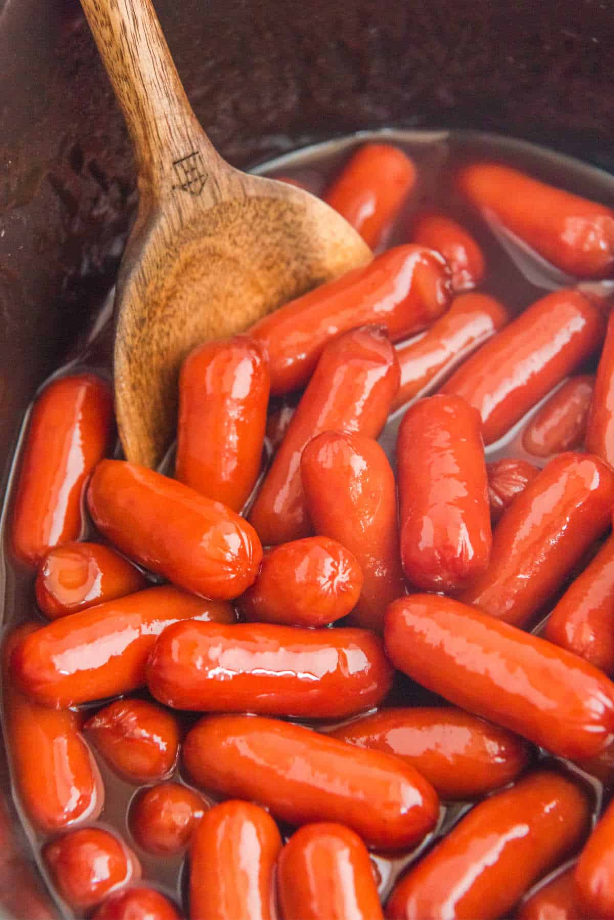 Little smokies in a slow cooker with a wooden spoon.