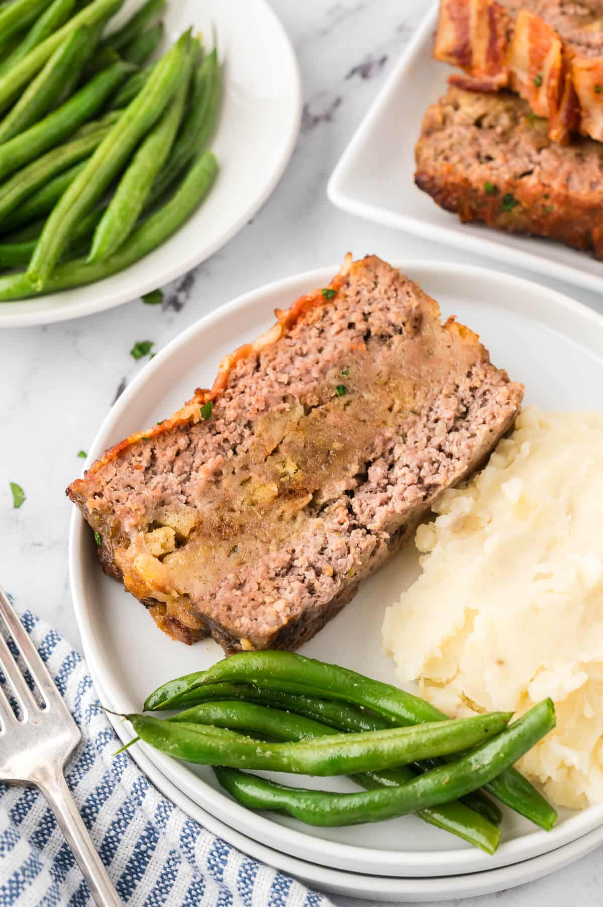 Sunday meatloaf slice on a plate with green beans and mashed potatoes.