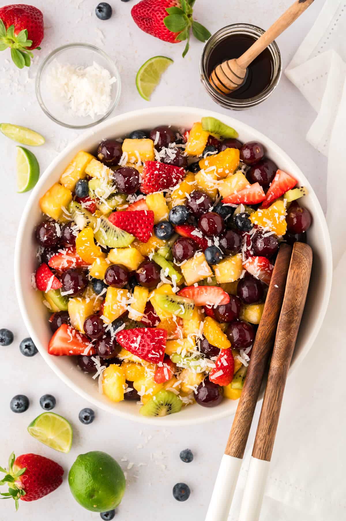 A bowl of rainbow fruit salad with wooden tongs.