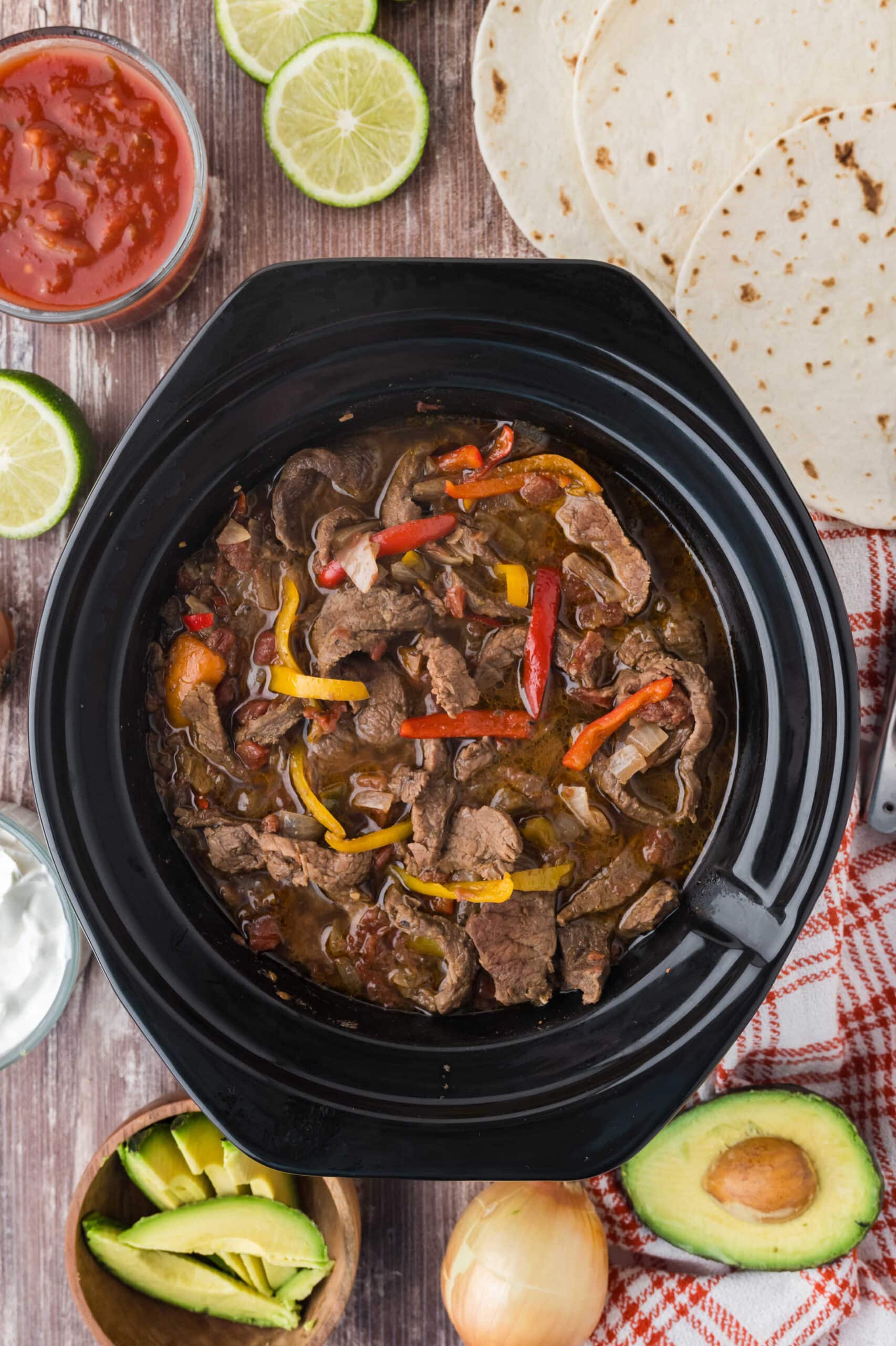 Steak fajitas in a slow cooker surrounded by toppings.