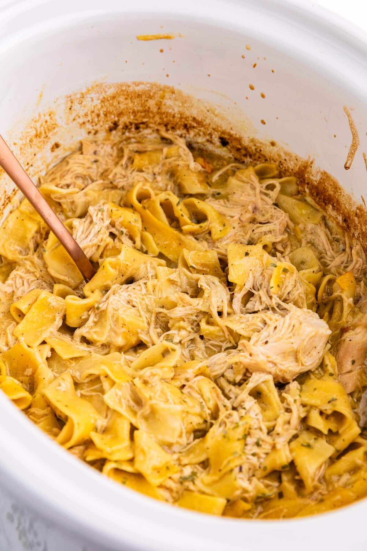 Slow cooker chicken and noodles in a white Crockpot and a serving spoon.
