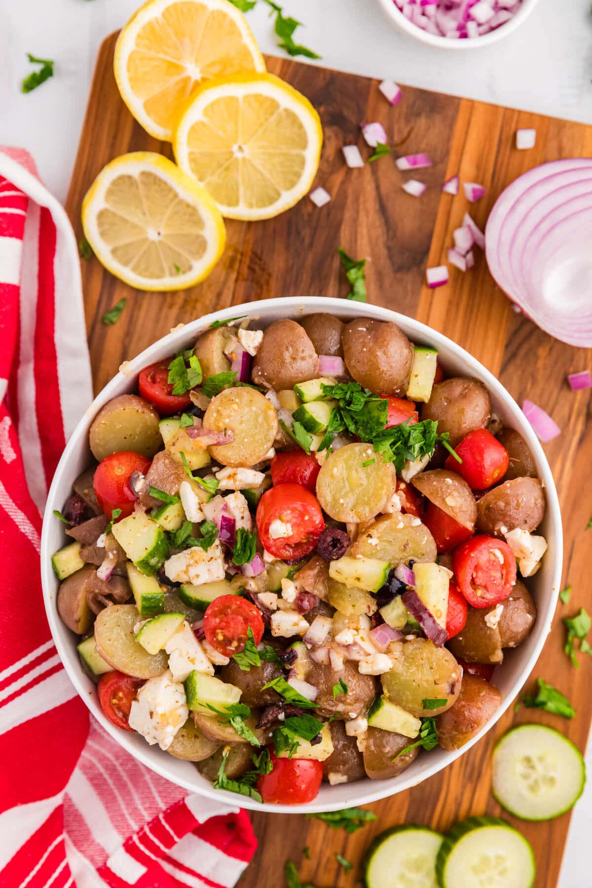 A bowl of Greek Potato Salad on a wooden surface surrounded by ingredients.