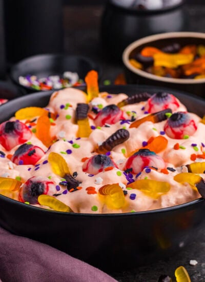 Wide hero shot of the recipe in a black bowl on a dark surface with scattered gummy worms and topping bowls behind.