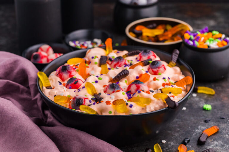 Wide hero shot of the recipe in a black bowl on a dark surface with scattered gummy worms and topping bowls behind.