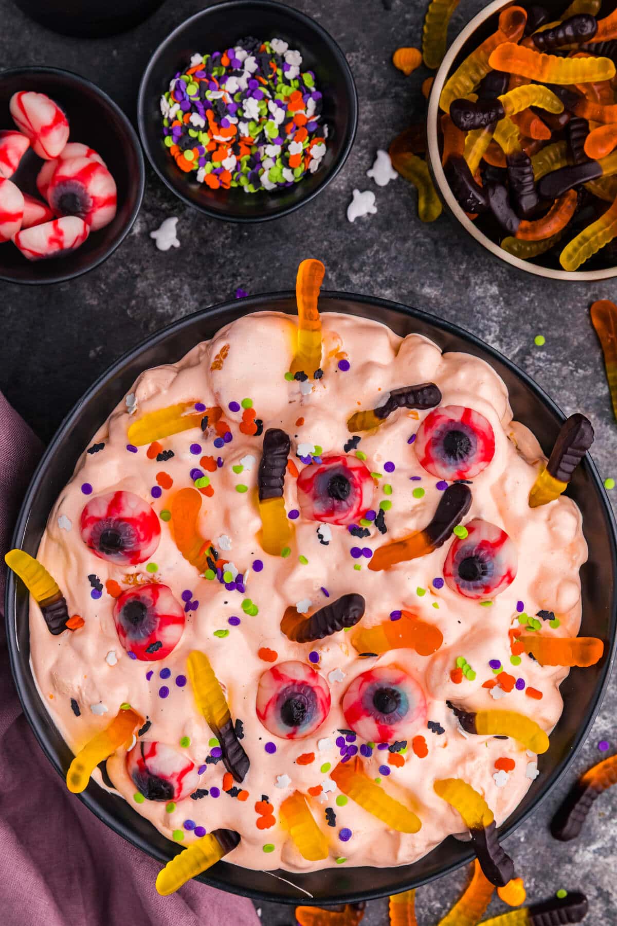 Overhead bowl of Halloween Fluff Salad tinted orange with gummy worms, candy eyeballs, and Halloween sprinkles.