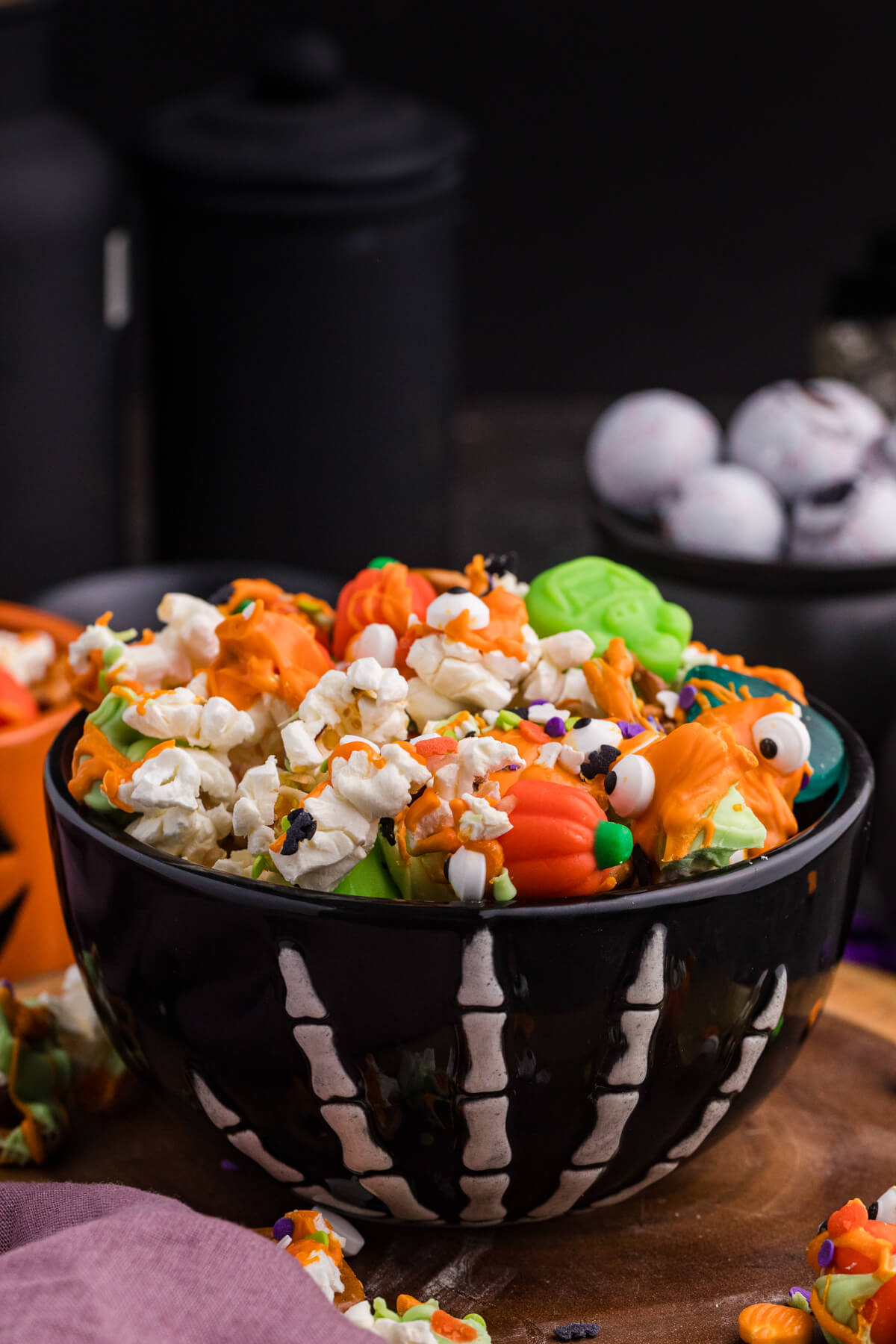 Bowl of the recipe on a wood board with orange and green drizzle, pretzels, mellowcreme pumpkins, candy eyeballs, and festive sprinkles.