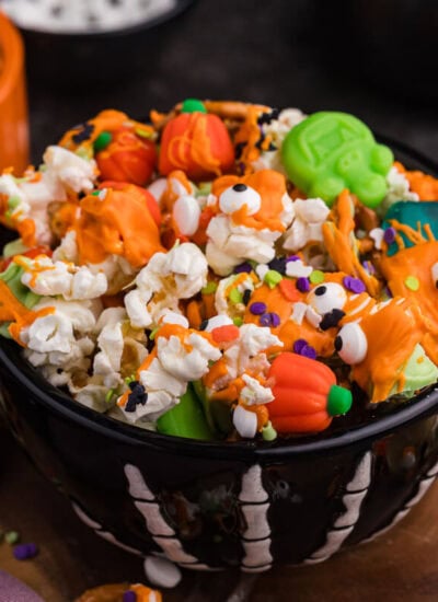 Black skeleton bowl filled with the recipe topped with orange and green candy melt drizzle, pretzels, mellowcreme pumpkins, and candy eyeballs.