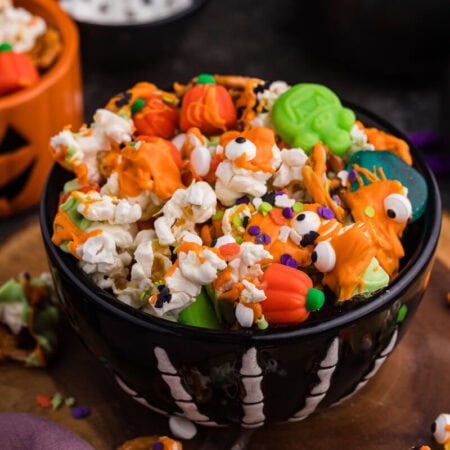 Black skeleton bowl filled with the recipe topped with orange and green candy melt drizzle, pretzels, mellowcreme pumpkins, and candy eyeballs.