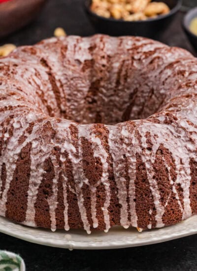 A full cake covered in a thin vanilla glaze sits on a white plate. Fresh apples, chopped walnuts, and a small bowl of applesauce are blurred in the background, with a green-striped cloth nearby.