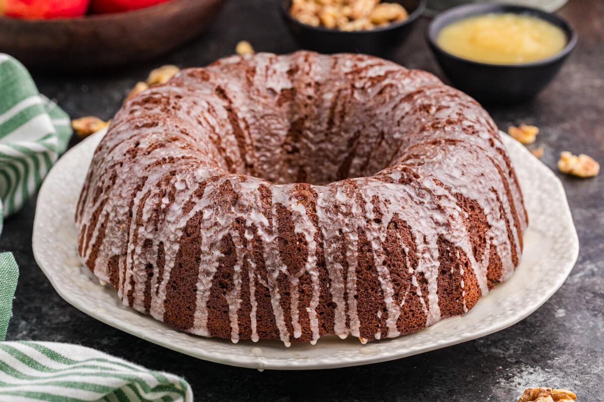 A full cake covered in a thin vanilla glaze sits on a white plate. Fresh apples, chopped walnuts, and a small bowl of applesauce are blurred in the background, with a green-striped cloth nearby.
