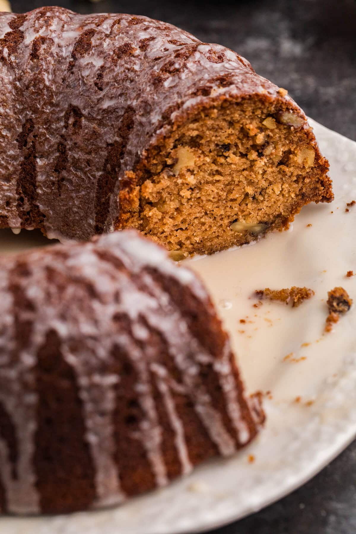 A close-up view of a sliced bundt cake, showing its moist, spiced interior with small walnut pieces and a light vanilla glaze dripping down the sides onto the plate.
