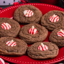 Chocolate cookies topped with red and white striped peppermint kisses, arranged in a circle on a red plate with festive decorations nearby.