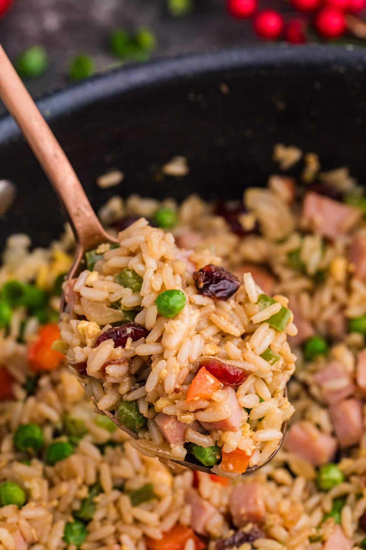 A close-up of a spoonful of fried rice with peas, carrots, ham, and cranberries held above a bowl of the same dish.