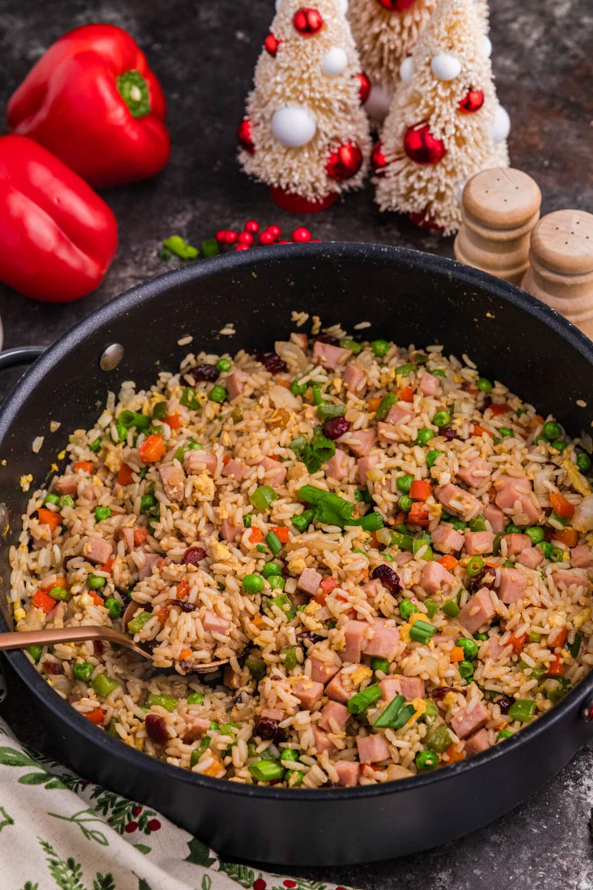 A skillet filled with Christmas Fried Rice sits on a dark countertop near red bell peppers, spice shakers, and holiday tree decorations.