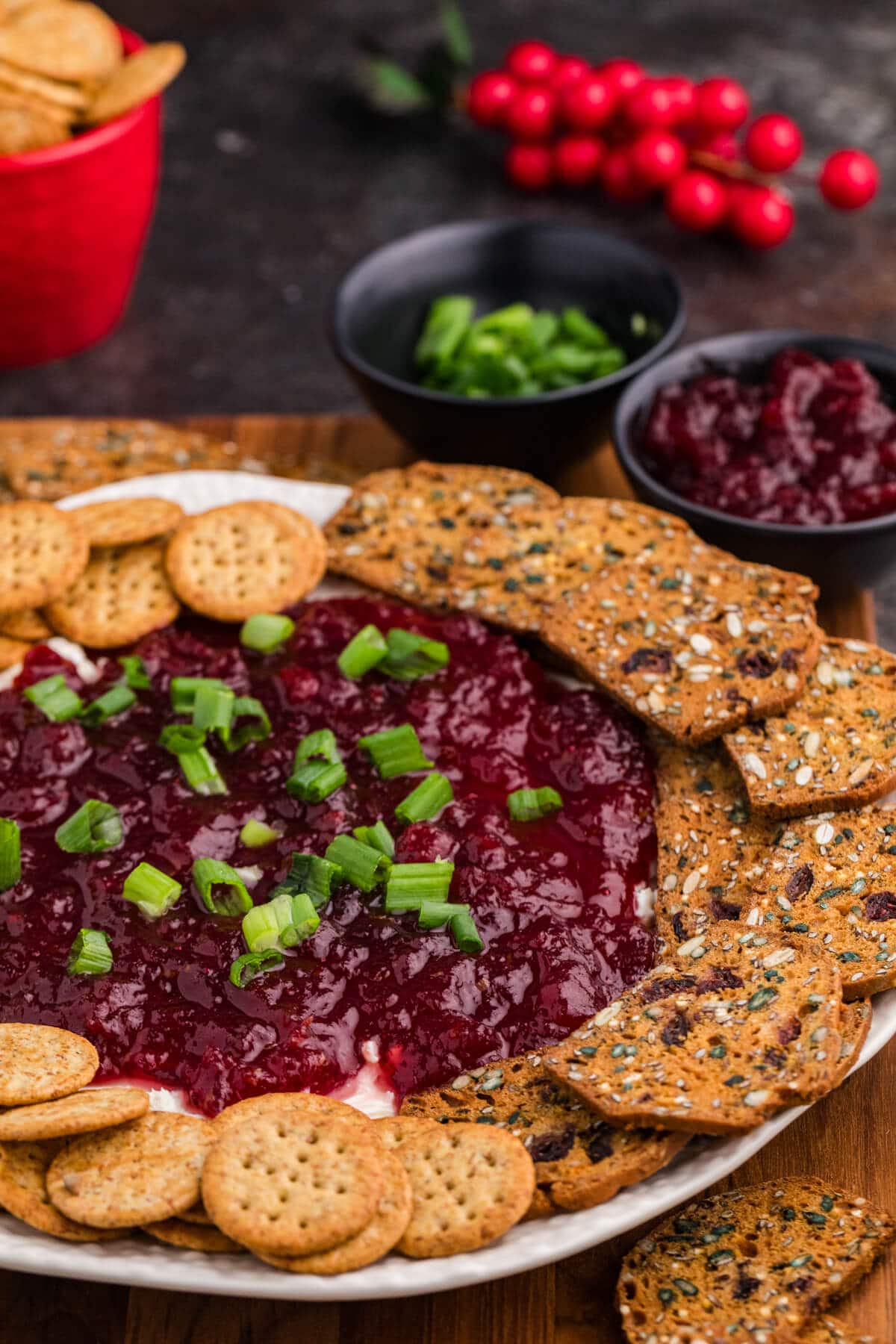 A platter with a vibrant cranberry dip topped with green onions, surrounded by assorted crackers. Small bowls of green garnish and cranberry sauce are in the background.