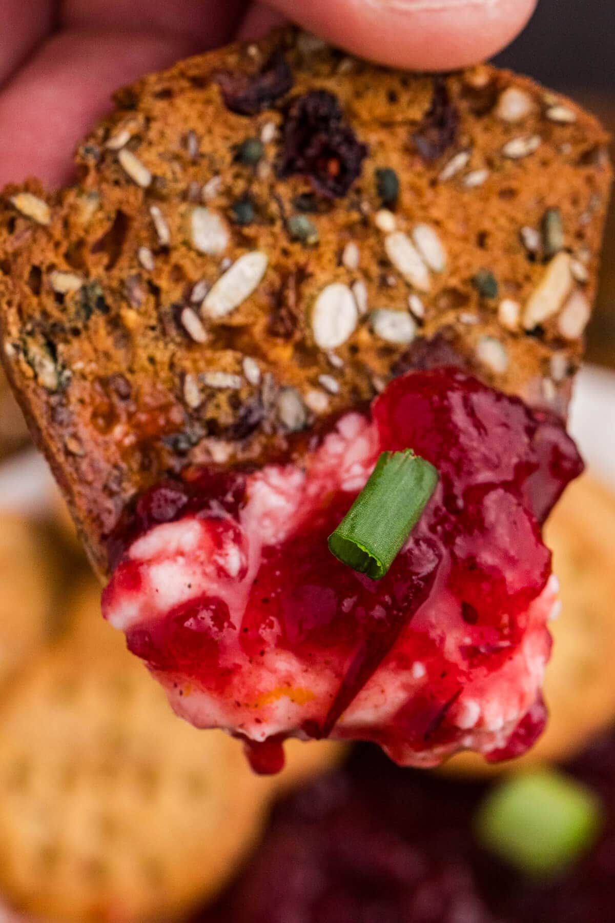 Close-up of a seeded cracker topped with creamy cheese, bright red cranberry sauce, and a green onion sliver, held by fingers.