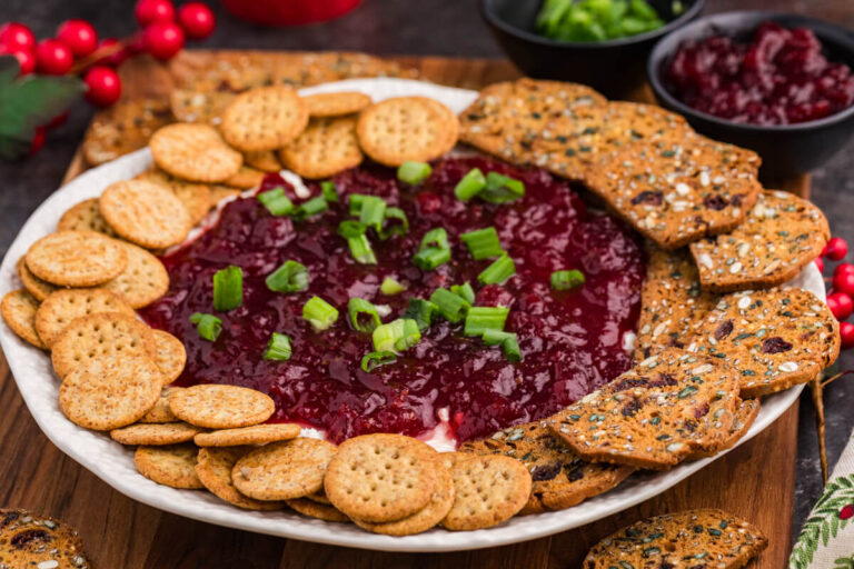 A platter of round crackers and multigrain crisps surrounds a dip topped with red cranberry sauce and chopped green onions, conveying a festive and inviting feel.