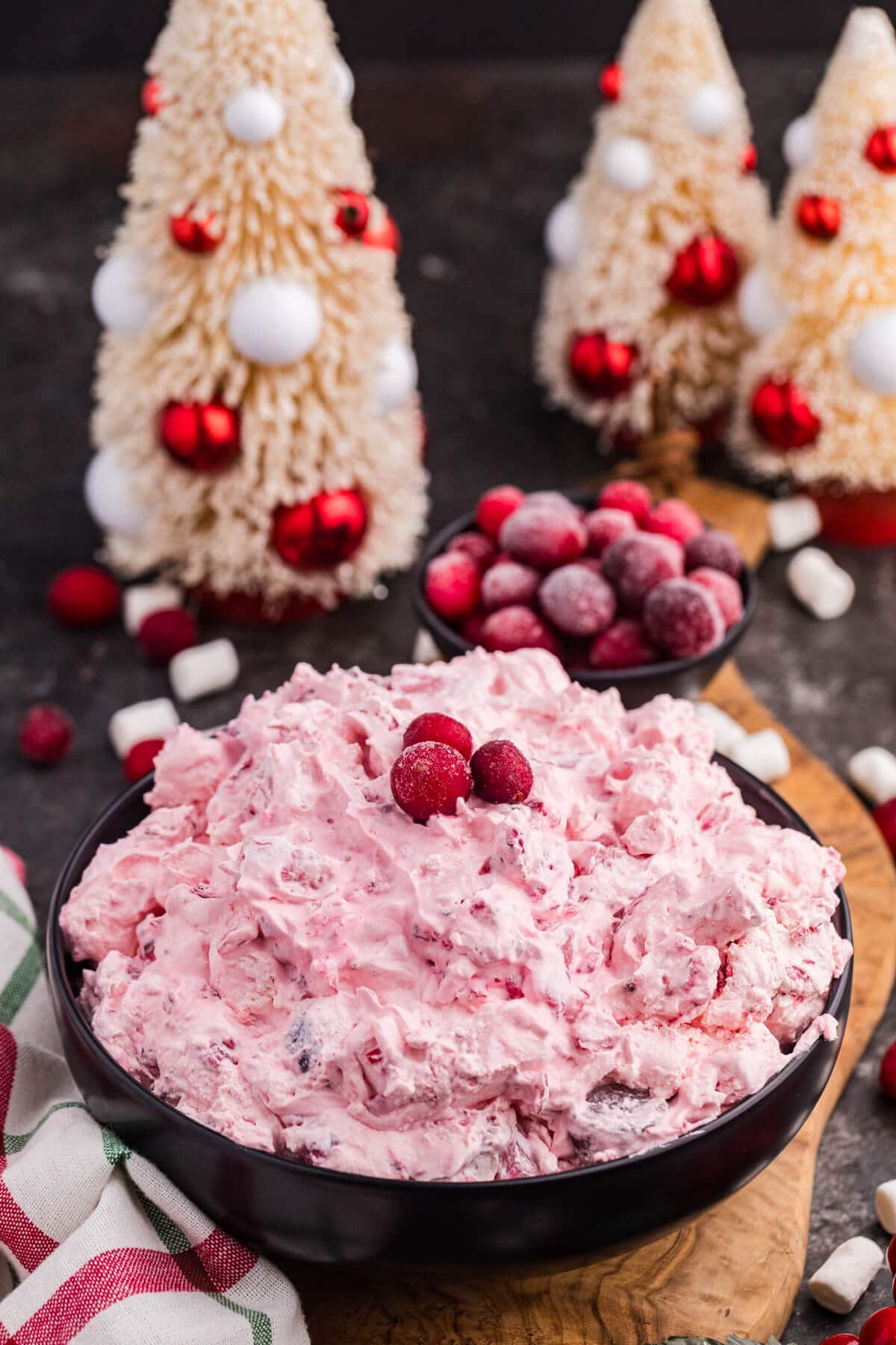 Cranberry fluff salad in a black bowl, topped with cranberries, surrounded by mini marshmallows and Christmas decor with festive trees and frosted berries.